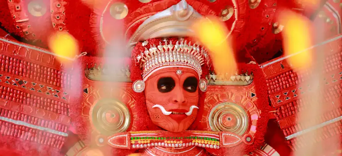 A Theyyam performer in red costume and painted face, seen through festival flames