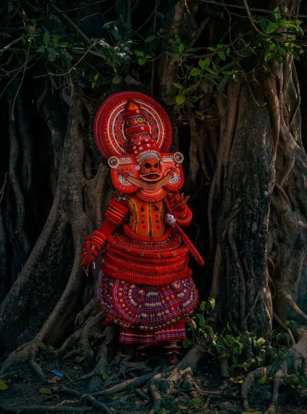 A Theyyam performer in ritual costume before the roots of a banyan tree, Kerala