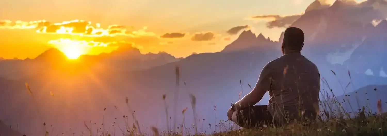 A man sitting on a grassy ridge at sunrise, looking out at distant mountain peaks