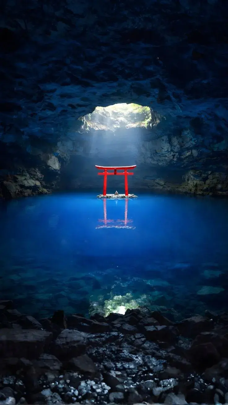 A red torii gate standing in luminous blue water inside a sea cave