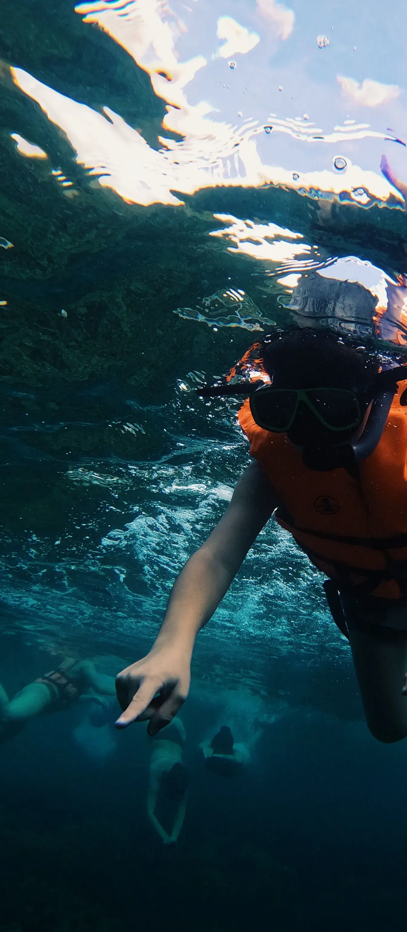 A snorkeller in an orange life jacket seen from under the water, the surface bright above