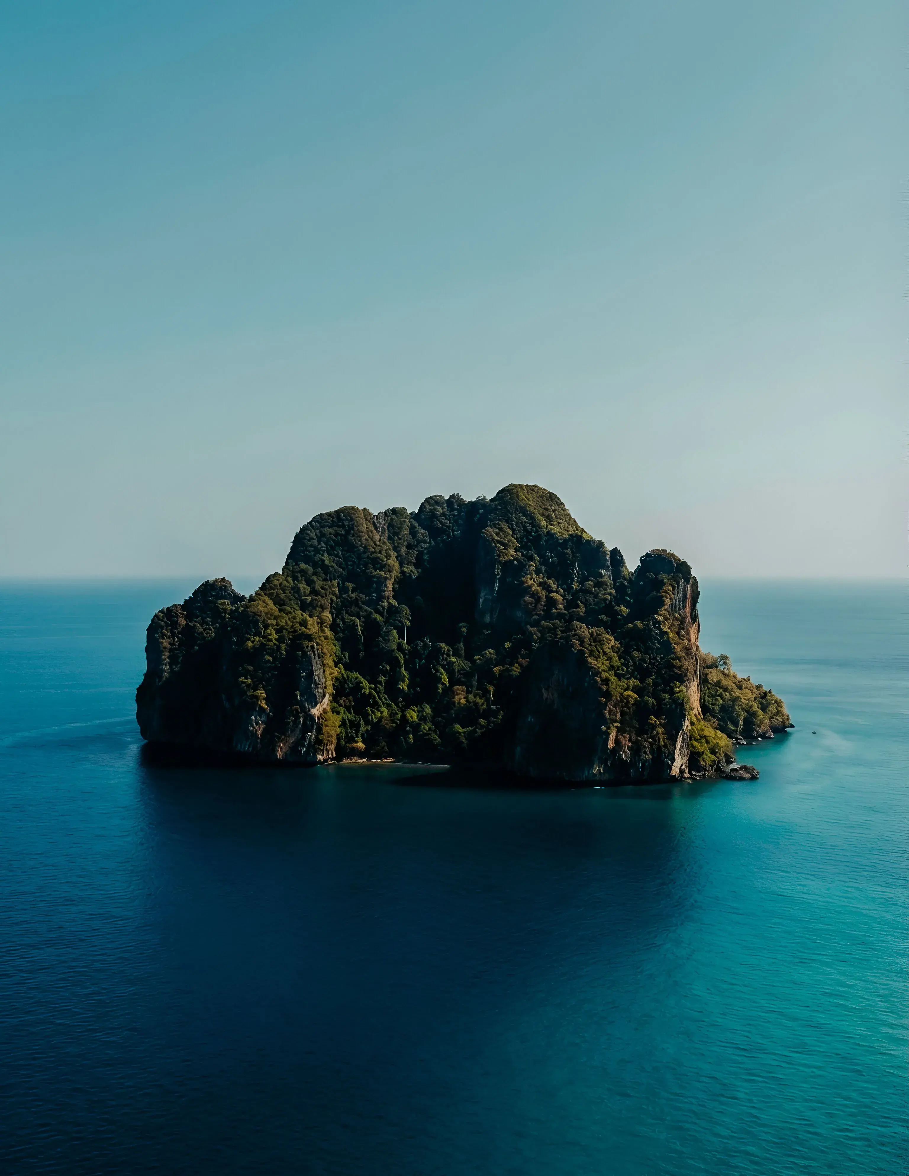 A rocky, wooded island alone in a calm turquoise sea under a pale sky