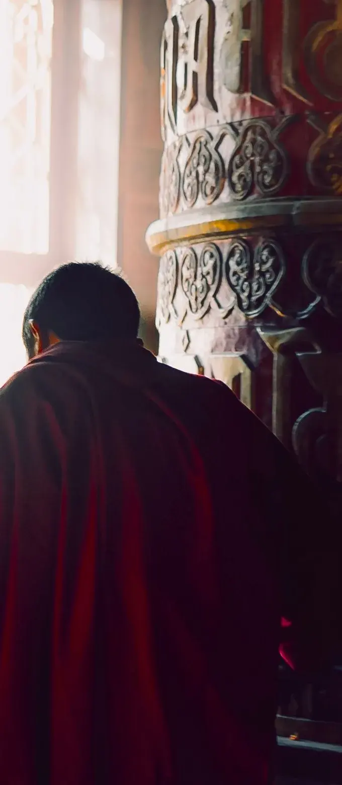 A monk in red robes beside a tall red-and-gold prayer wheel, light from a window behind him