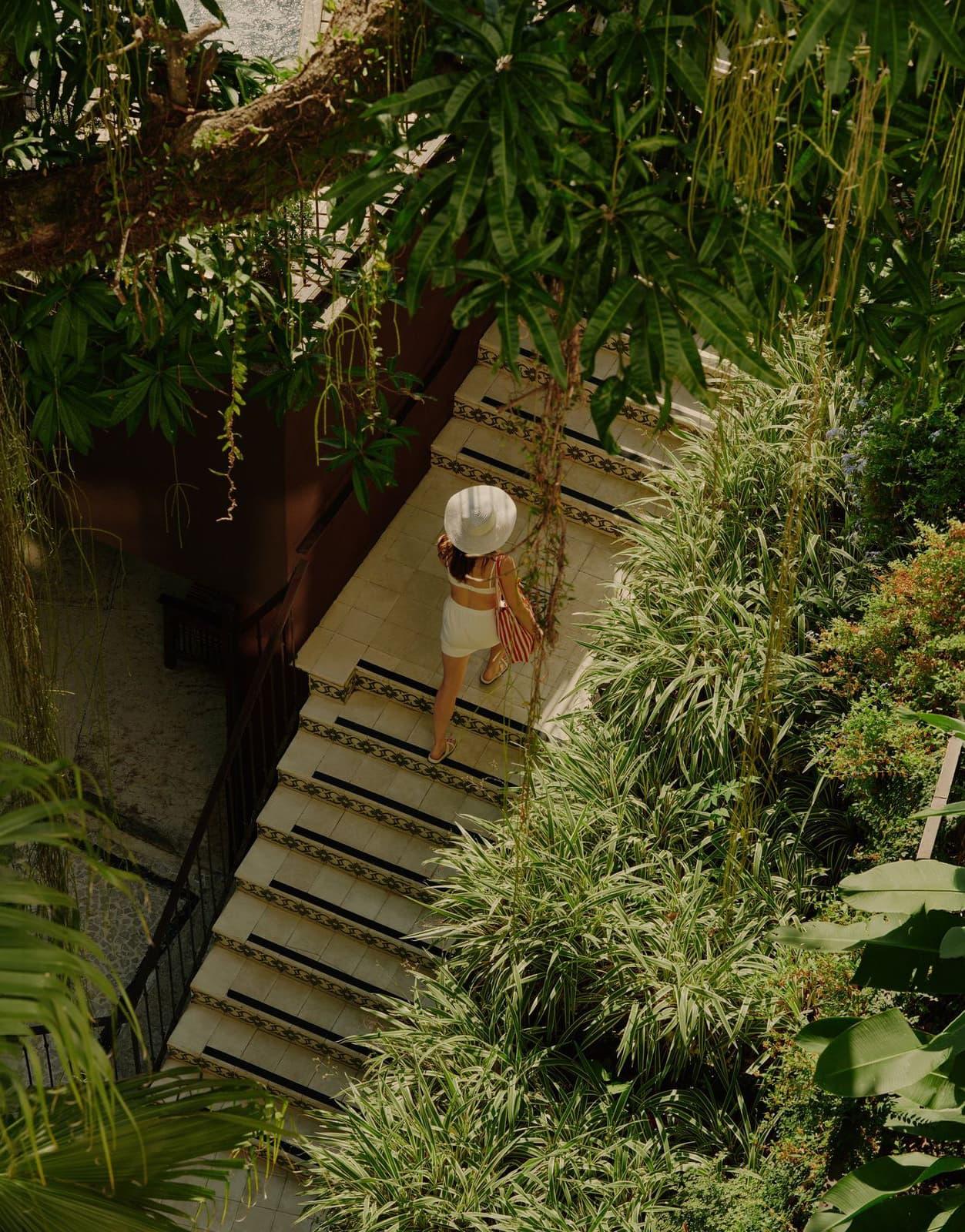 A woman in a white hat walking down tiled steps between tropical foliage, seen from above